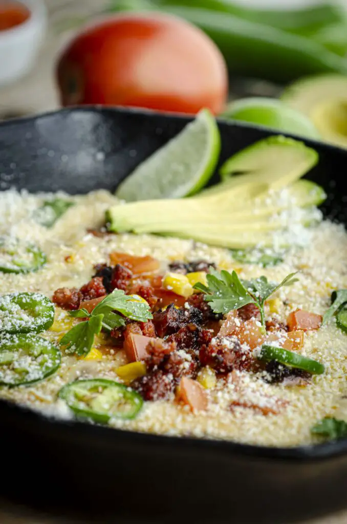 A closeup of a skillet prepared with queso fundido topped with garnishes of cooked chorizo, fresh cilantro, corn, chopped tomato, sliced avocado, a lime wedge, and sprinkled with cotija cheese. A fresh tomato and green chiles can be seen in the background. 