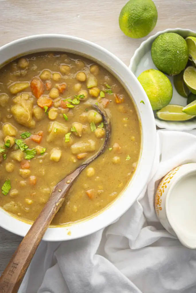 Looking down into a large white pot filled with coconut curry stew featuring chickpeas. A wooden ladel sits in the stew and the stew is surrounded with a cup of coconut cream and a container filled with limes.