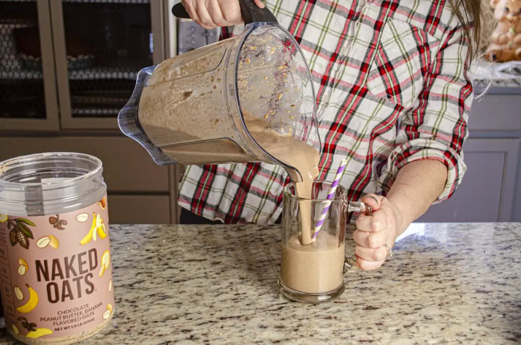 A lady pours a Chocolate Peanut Butter Banana Naked Oats smoothie from a blender into a glass mug with a spiral stripped straw. 