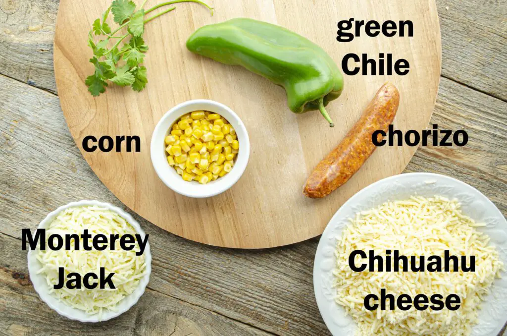 Looking down on a wooden tabletop where all the ingredients needed to make queso fundido are shown and labeled, including: chorizo, green chile, corn, queso chihuahua and Monterey Jack cheese.