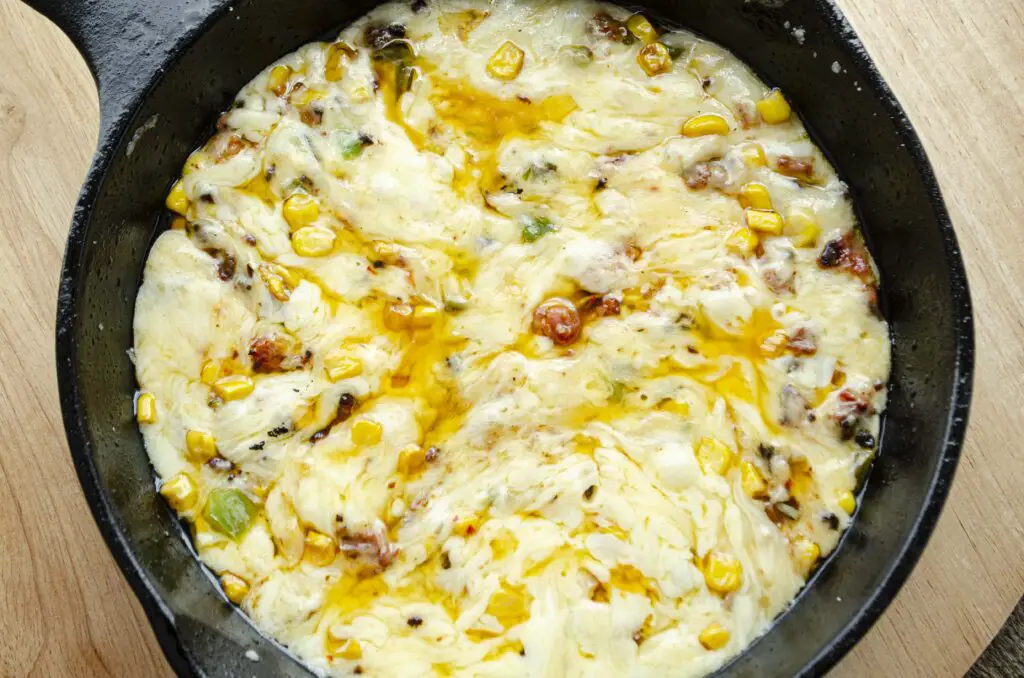 Looking down into a cast-iron skillet with melty white cheese mixed with chorizo, corn and chopped chile pepper with a little oil on top,