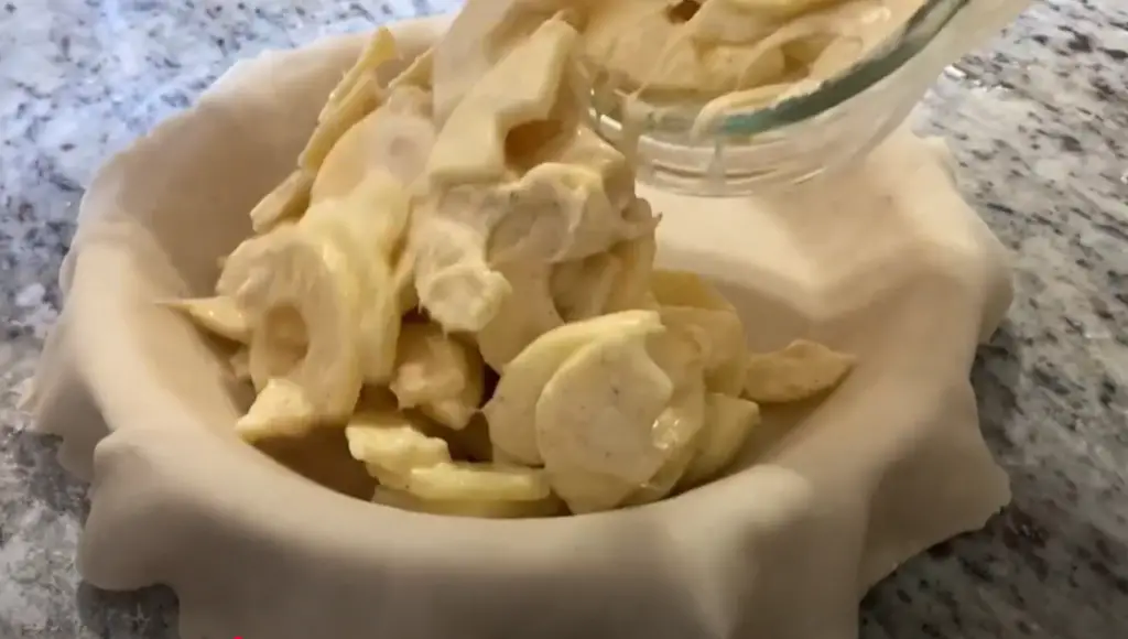 Apple pie filling is being poured into a prepared pie plate to make Browned Butter Cream Apple Pie with crumble topping.