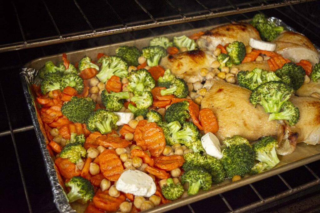 A sheet pan of Honey Garlic Chicken thighs roasts in the oven with sliced carrots and broccoli florets