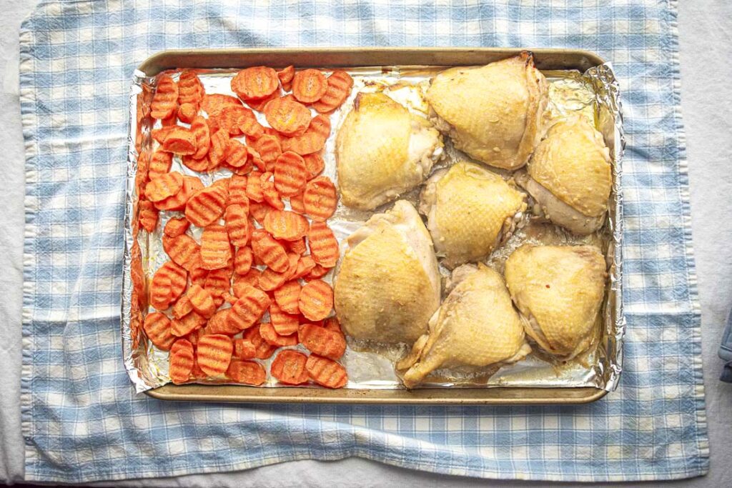 A metal sheet pan tray is loaded with sliced carrots on one side and chicken thighs (skin on, bone in) on the other side to make Honey Baked Chicken Thighs.