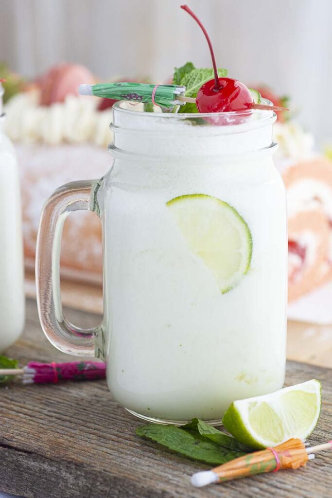 A glass mug filled with Brazilian 'Swiss Lemonade' is topped with garnishes and sits in front of a pink cake with fruit on top.