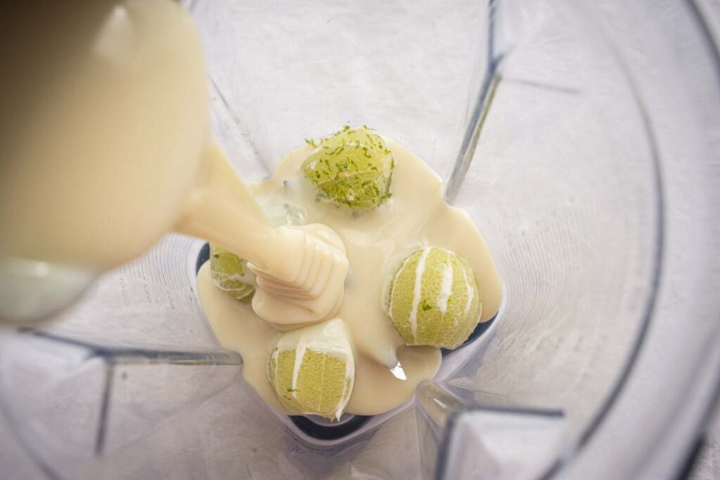 Looking down into a blender containing freshly peeled limes, lime zest, and sweetened condensed milk is poured over the top to make Brazilian Limeade.