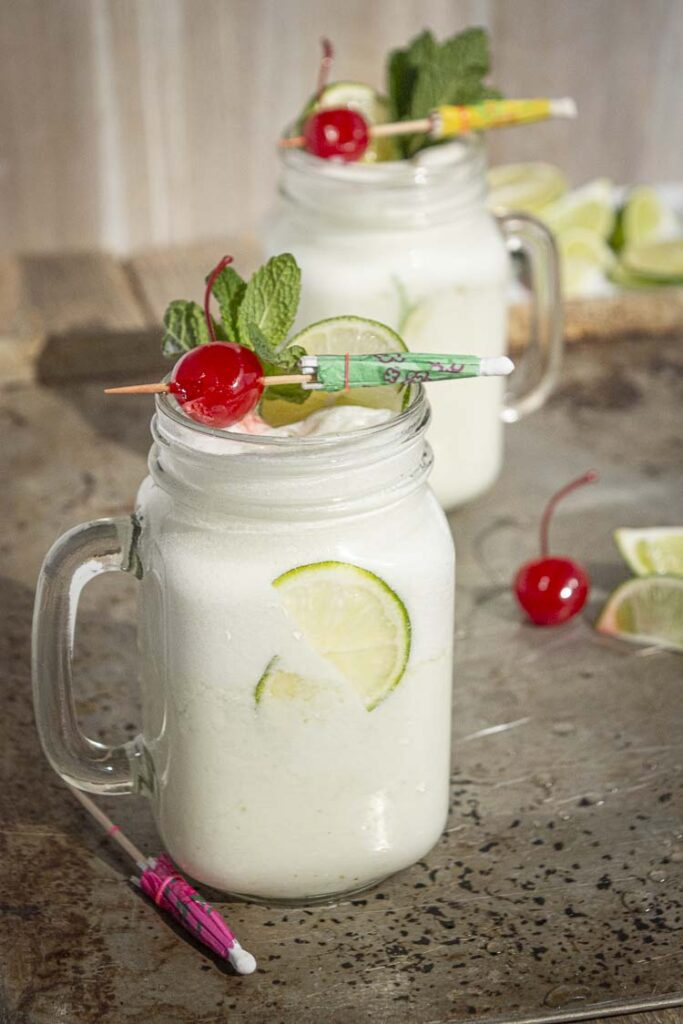 Two glass mason jar mugs filled with white Brazilian Limeade are topped with colorful garnishes.