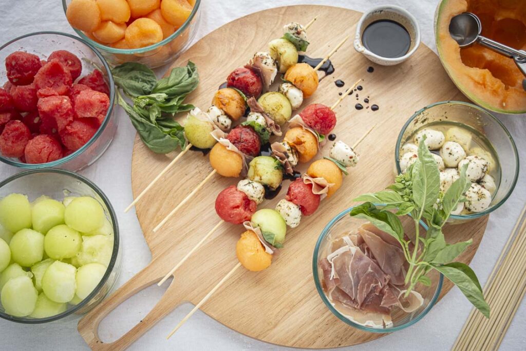 Looking down at a white tabletop spread with food. Shown in the center are four melon skewers garnished with a sprinkle of balsamic vinegar surrounded by various glass serving bowls full of different types of melon.
