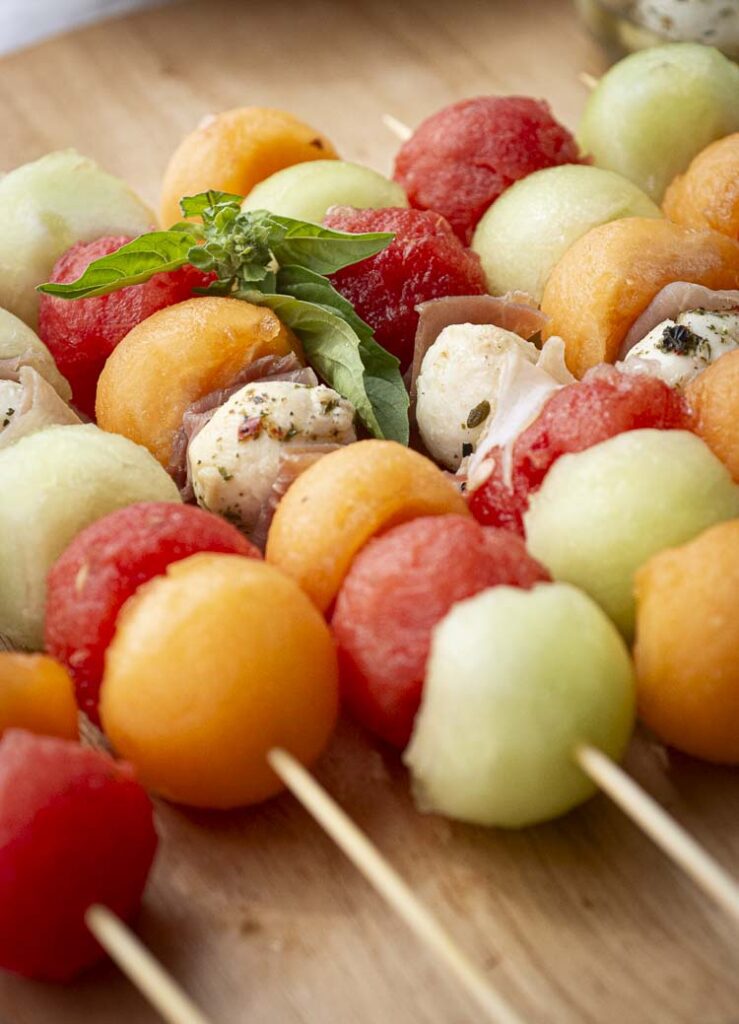 A closeup of melon skewers made with honeydew, watermelon, cantaloupe, mozzarella and prosciutto slices garnished with a sprig of fresh mint.
